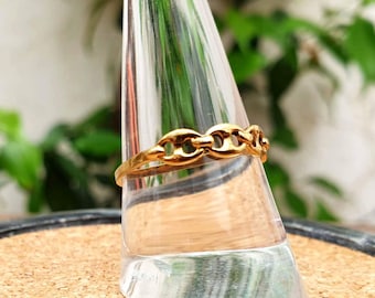 May include: A gold-colored ring with a chain link design. The ring is displayed on a clear glass object, with green foliage in the background. The ring's design features a series of interconnected oval shapes, creating a continuous chain-like pattern.