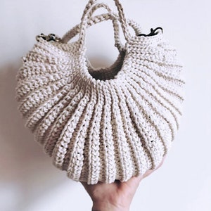 Peut inclure: Un sac en crochet de couleur crème avec une forme arrondie et deux anses. Le sac est tenu dans une main.