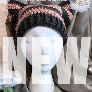 May include: A handmade cat ear beanie in dark gray and dusty rose stripes. The hat is displayed on a white mannequin head. The text "CAT EAR BEANIE" is at the top, and "NEW" is across the front. The Etsy shop name is at the bottom.