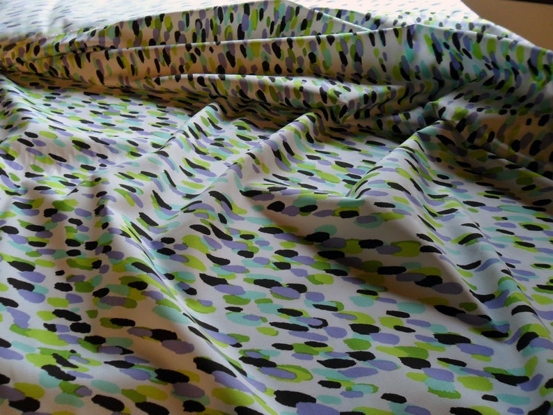 K&ouml;nnte beinhalten: Wei&szlig;er Stoff mit einem sich wiederholenden Muster aus abstrakten Formen in Schwarz, Gr&uuml;n, Hellblau und Lila. Der Stoff ist drapiert, zeigt Falten und Schatten und hat ein weiches, flie&szlig;endes Aussehen.