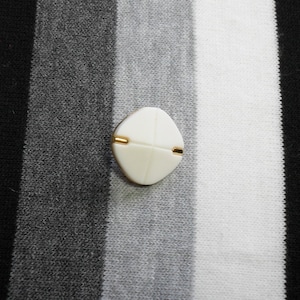 May include: A cream-colored, diamond-shaped button with gold-colored accents. The button is centered on a background of black, gray, and white vertical stripes. The button has a minimalist design.