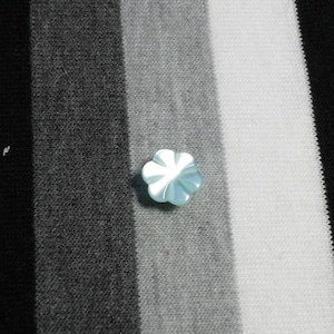 Puede incluir: Un pequeño botón en forma de flor azul claro con seis pétalos. El botón está hecho de un material translúcido y tiene un brillo ligeramente iridiscente. El botón está sobre un fondo de tela a rayas negras, grises y blancas.