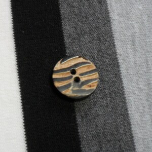 May include: A round button with two holes, featuring a brown and gray zebra stripe design. The button is set against a backdrop of black, gray, and white striped fabric, creating a contrasting visual.