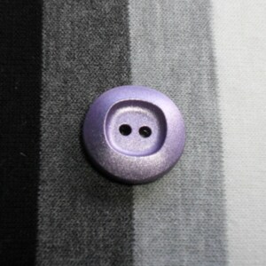 May include: A close-up of a round, lavender button with two small holes in the center. The button is set against a background of black, gray, and white striped fabric. The button has a slightly metallic sheen.
