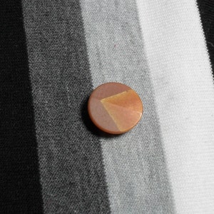 May include: A round, copper-colored button with a geometric design of triangles in shades of brown and gold. The button is set against a background of black, gray, and white striped fabric.