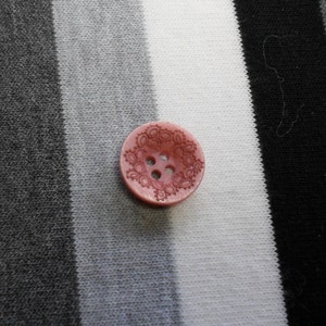 May include: A light pink button with a decorative floral pattern and four holes. The button is set against a backdrop of black, white, and gray striped fabric, creating a contrast in color and texture.