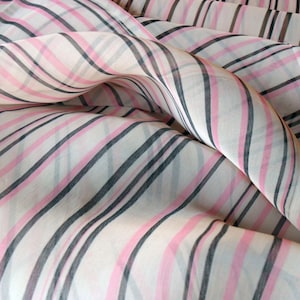 Pode incluir: Close-up de um tecido transparente com um padr&atilde;o listrado. O tecido apresenta listras alternadas rosa, pretas e cinzas sobre um fundo branco. O tecido &eacute; drapeado, criando dobras e curvas suaves.