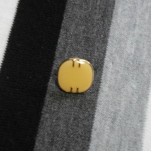 May include: A small, oval-shaped, yellow button with a gold-colored border. The button is set against a background of black, gray, and white striped fabric. The button has two vertical gold lines.