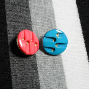 May include: Two round buttons, one coral and one turquoise, with a textured design. The buttons have a gold-colored rim and are set against a black, white, and gray striped background. These buttons are suitable for sewing projects.