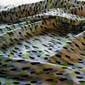 K&ouml;nnte beinhalten: Wei&szlig;er Stoff mit einem sich wiederholenden Muster aus bunten, unregelm&auml;&szlig;igen Formen. Das Design zeigt schwarze, gr&uuml;ne, hellblaue und violette Pinselstriche, die &uuml;ber die Oberfl&auml;che verstreut sind. Der Stoff ist gefaltet und erzeugt Schatten und Textur.