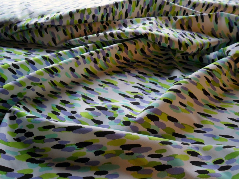 K&ouml;nnte beinhalten: Wei&szlig;er Stoff mit einem sich wiederholenden Muster aus bunten, unregelm&auml;&szlig;igen Formen. Das Design zeigt schwarze, gr&uuml;ne, hellblaue und violette Pinselstriche, die &uuml;ber die Oberfl&auml;che verstreut sind. Der Stoff ist gefaltet und erzeugt Schatten und Textur.