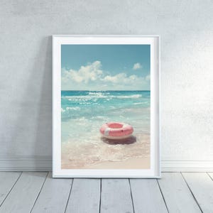 Puede incluir: Impresión artística enmarcada que representa una escena de playa serena. Un flotador inflable rosa y blanco flota en las suaves olas del océano turquesa. El cielo es azul claro con nubes blancas, creando un ambiente veraniego tranquilo.