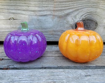 Puede incluir: Dos calabazas decorativas, una morada con purpurina y un tallo verde, la otra naranja con un tallo marrón. Las calabazas están sobre una superficie de madera envejecida. Estas calabazas son probablemente para la decoración de otoño o Halloween.