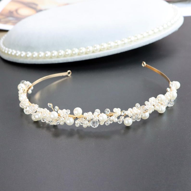 May include: A delicate gold-toned bridal headband adorned with faux pearls and clear crystals. The tiara-style headpiece features a flexible wire frame and is designed for special occasions. The background includes a white, pearl-trimmed plate.