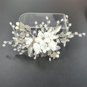 Op de afbeelding: Een bruidskam met witte stoffen bloemen, zilveren bladeren en sprankelende strass-steentjes. De kam is versierd met imitatieparels en delicate, met kralen bezette takjes, waardoor een elegant accessoire ontstaat voor bruiloften of speciale gelegenheden.