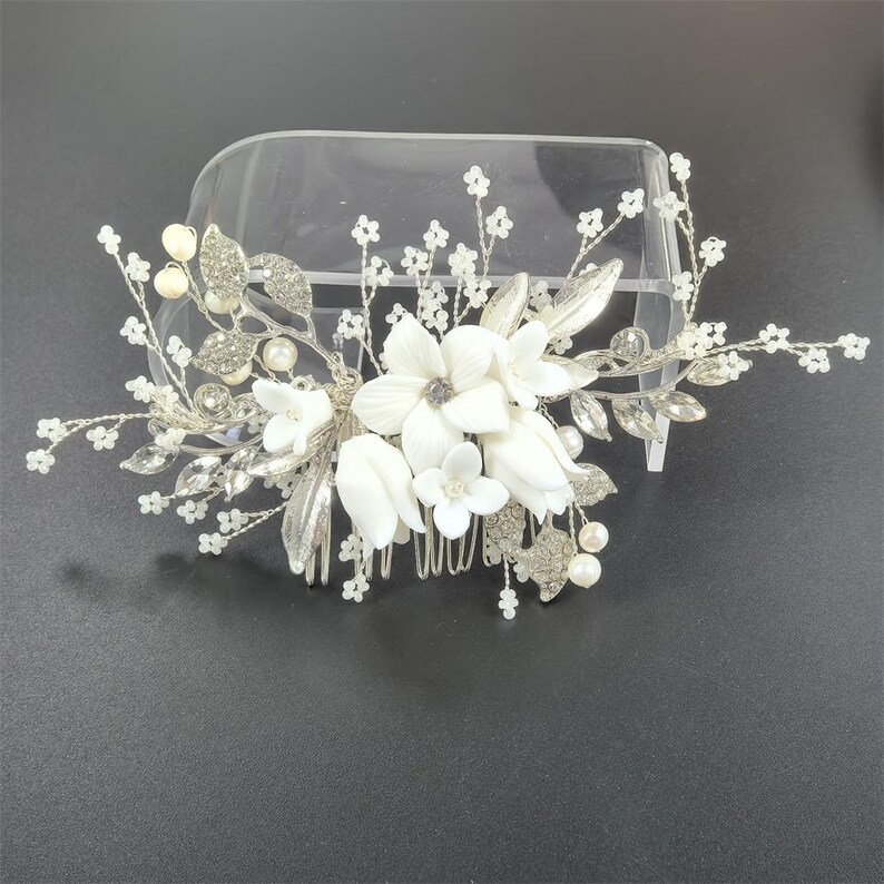 Op de afbeelding: Een bruidskam met witte stoffen bloemen, zilveren bladeren en sprankelende strass-steentjes. De kam is versierd met imitatieparels en delicate, met kralen bezette takjes, waardoor een elegant accessoire ontstaat voor bruiloften of speciale gelegenheden.