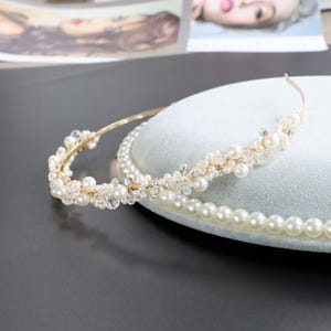 May include: A gold-toned bridal headband with faux pearls and crystals. The delicate design features a thin band adorned with clusters of pearls and sparkling accents. A strand of pearls is also visible.