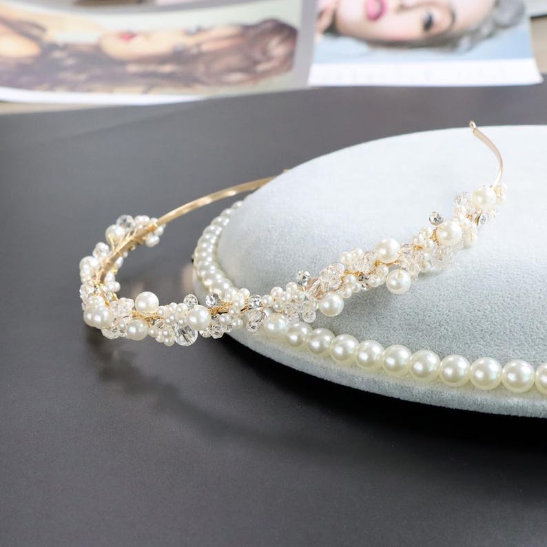 May include: A gold-toned bridal headband with faux pearls and crystals. The delicate design features a thin band adorned with clusters of pearls and sparkling accents. A strand of pearls is also visible.