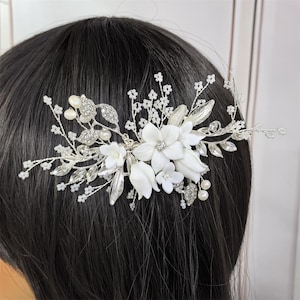 Op de afbeelding: Zilveren bruidskam met witte porseleinen bloemen, imitatieparels en sprankelende kristallen. Het bloemendesign bevat bladeren en kralenaccenten, waardoor een elegant accessoire ontstaat voor bruiloften of speciale gelegenheden.