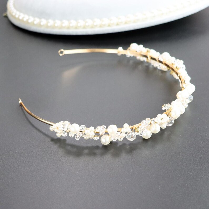 May include: A delicate gold-toned bridal headband. The headband is adorned with clusters of white pearls and clear crystals. The design is elegant and suitable for weddings or special occasions. The headband is curved and has a simple, refined aesthetic.