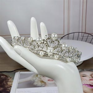 Pu&ograve; includere: Una tiara color argento con perle finte e strass trasparenti. La corona &egrave; tenuta da un espositore a forma di mano bianca. La tiara presenta un design floreale con molteplici accenti di perle. Lo sfondo include una superficie bianca e una rivista.