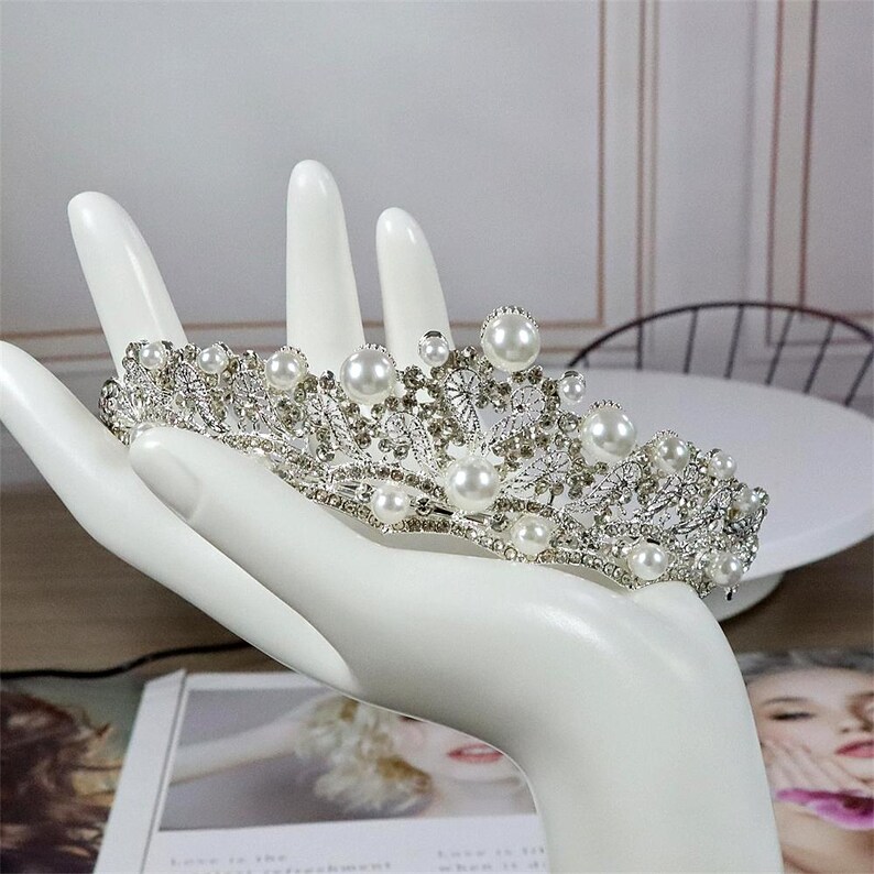 Pu&ograve; includere: Una tiara color argento con perle finte e strass trasparenti. La corona &egrave; tenuta da un espositore a forma di mano bianca. La tiara presenta un design floreale con molteplici accenti di perle. Lo sfondo include una superficie bianca e una rivista.