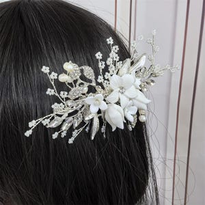 Op de afbeelding: Een zilverkleurige haar kam met witte bloemaccenten, imitatieparels en heldere kristallen. De kam is ontworpen met delicate bladeren en takjes, waardoor een elegant accessoire met bruidsthema ontstaat.