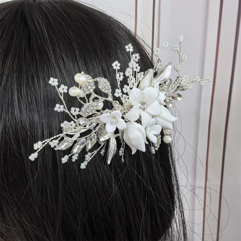Op de afbeelding: Een zilverkleurige haar kam met witte bloemaccenten, imitatieparels en heldere kristallen. De kam is ontworpen met delicate bladeren en takjes, waardoor een elegant accessoire met bruidsthema ontstaat.