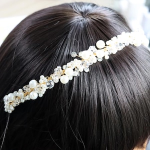 May include: A delicate, gold-toned headband adorned with clusters of white pearls and clear crystals. The bridal or special occasion hair accessory is set against a backdrop of dark hair, highlighting its intricate design.
