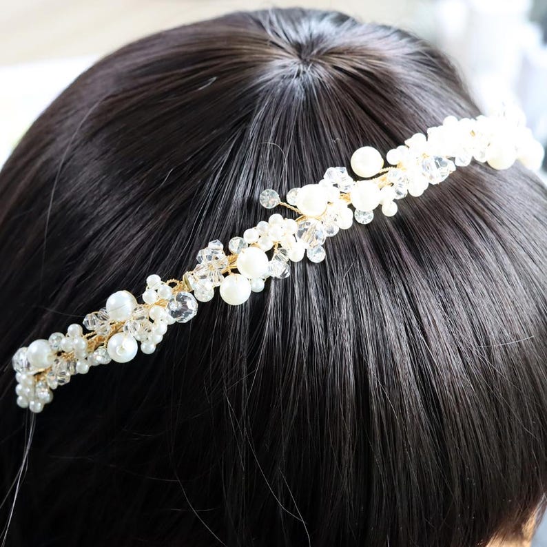 May include: A delicate, gold-toned headband adorned with clusters of white pearls and clear crystals. The bridal or special occasion hair accessory is set against a backdrop of dark hair, highlighting its intricate design.