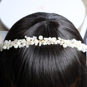 May include: A delicate, handcrafted bridal headband featuring a gold wire base adorned with clusters of white pearls and clear crystal beads. The headpiece is designed to sit atop dark hair, creating a shimmering, elegant effect. Perfect for weddings or special occasions.