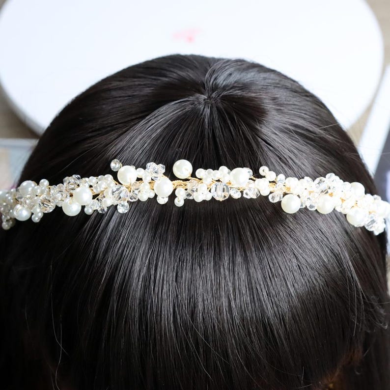 May include: A delicate, handcrafted bridal headband featuring a gold wire base adorned with clusters of white pearls and clear crystal beads. The headpiece is designed to sit atop dark hair, creating a shimmering, elegant effect. Perfect for weddings or special occasions.