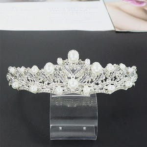 Pu&ograve; includere: Una tiara color argento ornata di perle finte e strass trasparenti. L'ornata corona presenta un delicato design traforato con una disposizione simmetrica di perle e accenti scintillanti. La tiara &egrave; esposta su un supporto trasparente.