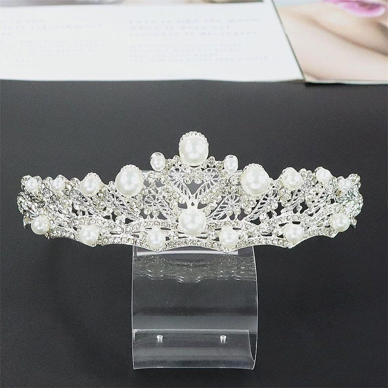Pu&ograve; includere: Una tiara color argento ornata di perle finte e strass trasparenti. L'ornata corona presenta un delicato design traforato con una disposizione simmetrica di perle e accenti scintillanti. La tiara &egrave; esposta su un supporto trasparente.
