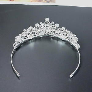 Pu&ograve; includere: Una tiara color argento con un motivo floreale decorativo e accenti di cristallo trasparente. La tiara ha una fascia curva ed &egrave; progettata per essere indossata sulla testa. I cristalli sono rotondi e incastonati al centro e sulla parte superiore della tiara.