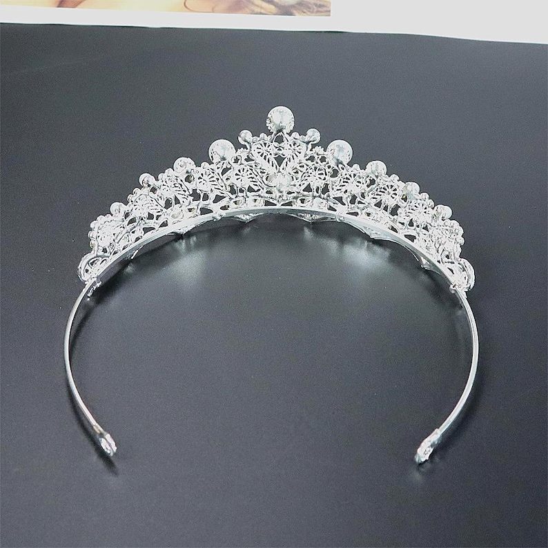 Pu&ograve; includere: Una tiara color argento con un motivo floreale decorativo e accenti di cristallo trasparente. La tiara ha una fascia curva ed &egrave; progettata per essere indossata sulla testa. I cristalli sono rotondi e incastonati al centro e sulla parte superiore della tiara.