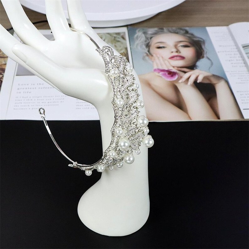 Pu&ograve; includere: Un copricapo da sposa argentato con un design curvo, adornato con perle finte e cristalli trasparenti. Il copricapo &egrave; esposto su un espositore per gioielli a forma di mano bianca, con una rivista sullo sfondo.