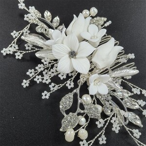 Op de afbeelding: Een zilverkleurige bruidskam met witte bloemen, imitatieparels en sprankelende strass-steentjes. De kam heeft een delicaat ontwerp met meerdere bloemvormen en blad details, perfect voor bruiloften of speciale gelegenheden.