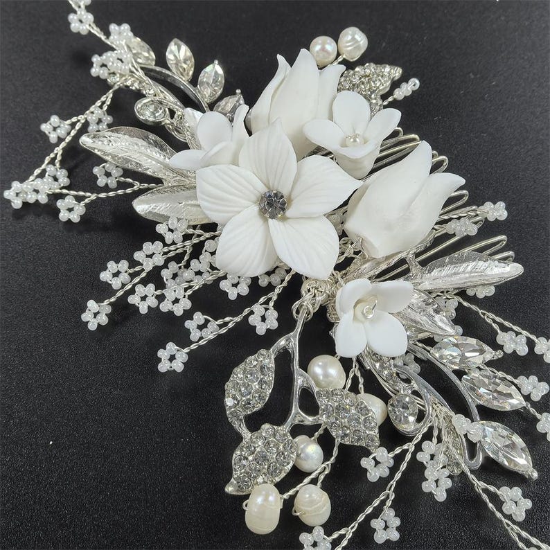 Op de afbeelding: Een zilverkleurige bruidskam met witte bloemen, imitatieparels en sprankelende strass-steentjes. De kam heeft een delicaat ontwerp met meerdere bloemvormen en blad details, perfect voor bruiloften of speciale gelegenheden.