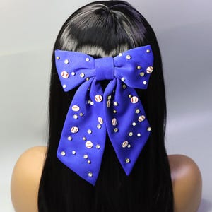 Peut inclure: Un grand nœud pour les cheveux bleu roi orné de petits ballons de baseball et de strass étincelants. Le nœud est noué de manière classique et se détache sur une chevelure noire. Les ballons de baseball sont blancs avec des coutures rouges.