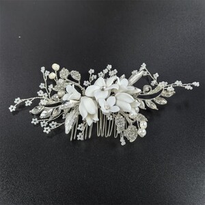 Op de afbeelding: Zilveren bruidskam met witte stoffen bloemen, imitatieparels en strass. De kam heeft een bloemenontwerp met bladeren en delicate kralenaccenten, ideaal voor bruiloften of speciale gelegenheden.