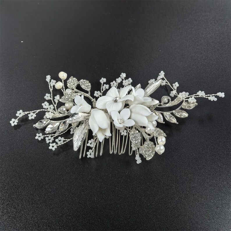 Op de afbeelding: Zilveren bruidskam met witte stoffen bloemen, imitatieparels en strass. De kam heeft een bloemenontwerp met bladeren en delicate kralenaccenten, ideaal voor bruiloften of speciale gelegenheden.