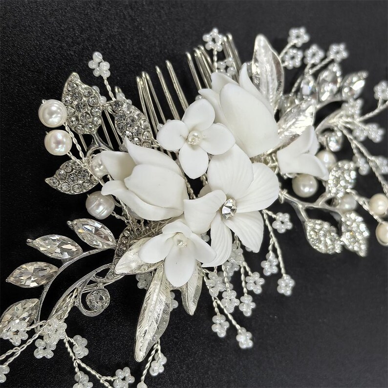 Op de afbeelding: Een zilverkleurige bruidskam met witte bloemaccenten, imitatieparels en sprankelende strass-steentjes. De kam heeft een delicaat ontwerp met meerdere bloemvormen en bladdetails, perfect voor bruiloften of speciale gelegenheden.