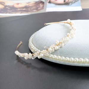 May include: A gold-toned bridal headband with faux pearls and clear crystals. The headband features a delicate design with a mix of small and large pearls, and sparkling crystals. The accessory is designed for weddings and special occasions.