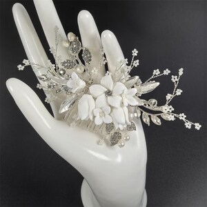 Op de afbeelding: Een bruidskam met witte porseleinen bloemen, zilveren bladeren en sprankelende kristallen. De kam is versierd met imitatieparels en delicate takjes, perfect voor een bruiloft of speciale gelegenheid. De kam is ontworpen om in het haar te worden gedragen.
