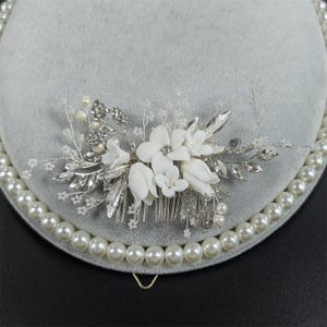 Op de afbeelding: Een bruidskam met witte stoffen bloemen, zilveren bladeren en sprankelende kristallen. De kam is versierd met imitatieparels en staat tegen een grijze achtergrond. Dit elegante haaraccessoire is perfect voor bruiloften en speciale gelegenheden.
