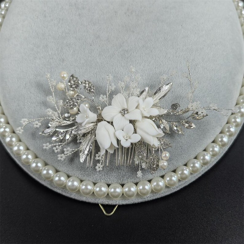 Op de afbeelding: Een bruidskam met witte stoffen bloemen, zilveren bladeren en sprankelende kristallen. De kam is versierd met imitatieparels en staat tegen een grijze achtergrond. Dit elegante haaraccessoire is perfect voor bruiloften en speciale gelegenheden.