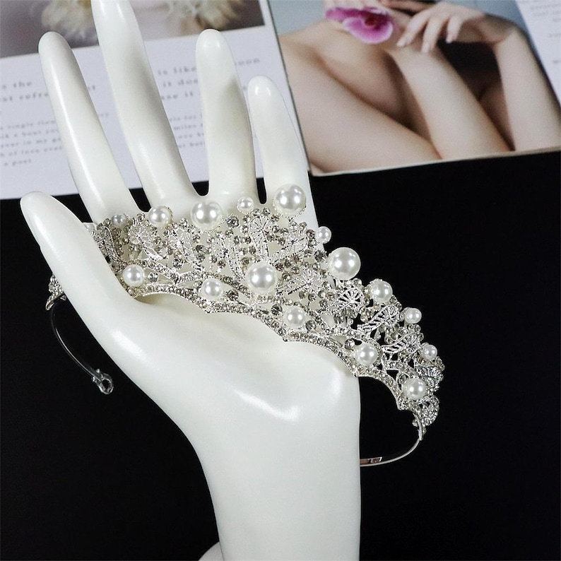 Pu&ograve; includere: Una tiara argentata con perle finte e strass trasparenti. La corona nuziale presenta un design delicato con accenti floreali e a foglia. La tiara &egrave; esposta su un espositore a forma di mano bianca.