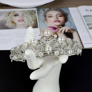 Pu&ograve; includere: Tiara argentata con perle finte e strass trasparenti. La corona &egrave; esposta su un supporto bianco a forma di mano, con una rivista sullo sfondo. La tiara presenta un design traforato e complesso, ideale per matrimoni o eventi speciali.