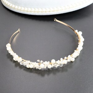 May include: A gold-toned metal headband adorned with white pearls and clear crystals. The delicate design features a curved shape, with the embellishments clustered along the front. The background is a dark gray surface.